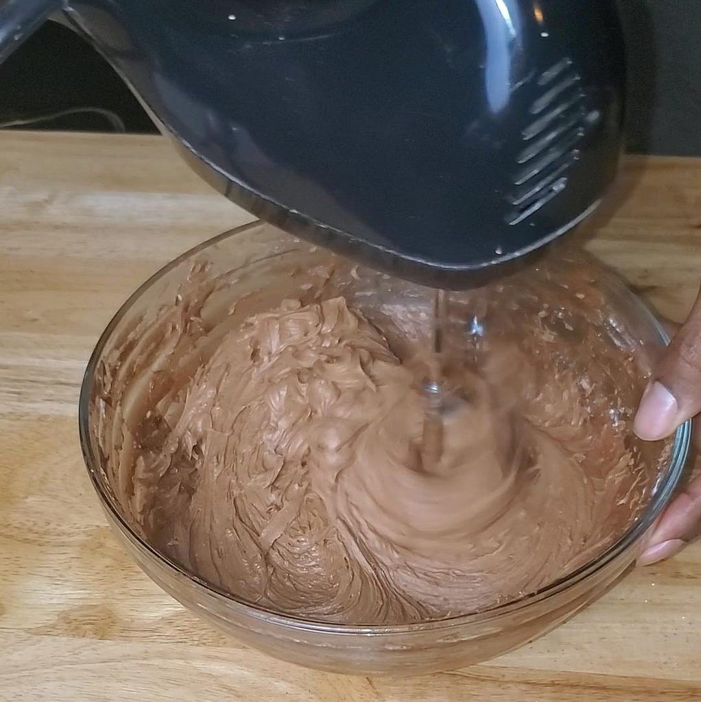 This image depicts chocolate cream cheese frositng being mixed together using a black hand mixer inside of a glass bowl.