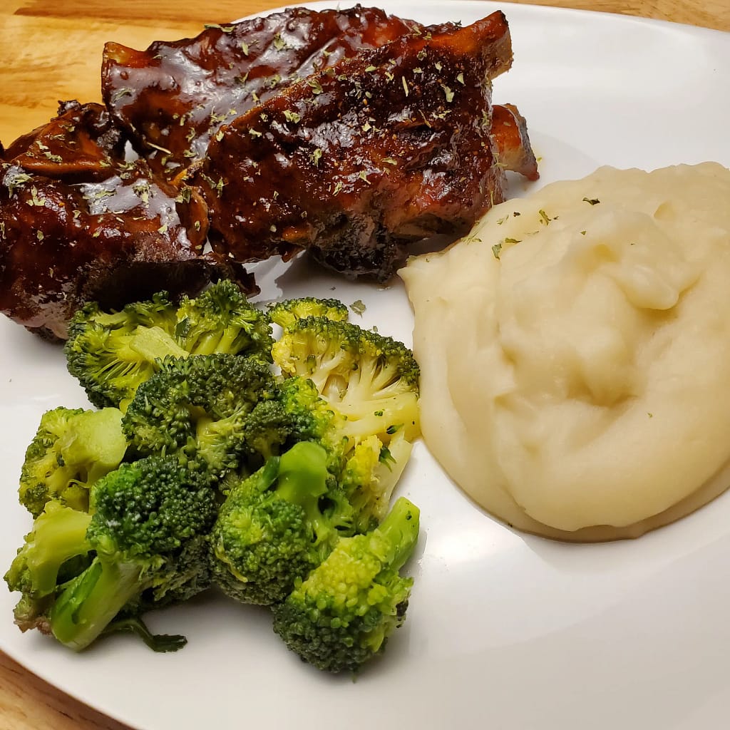 This image depicts Oven-roasted barbecue Pork Neck Bones on a white plate along side mashed potatoes and steamed broccoli.