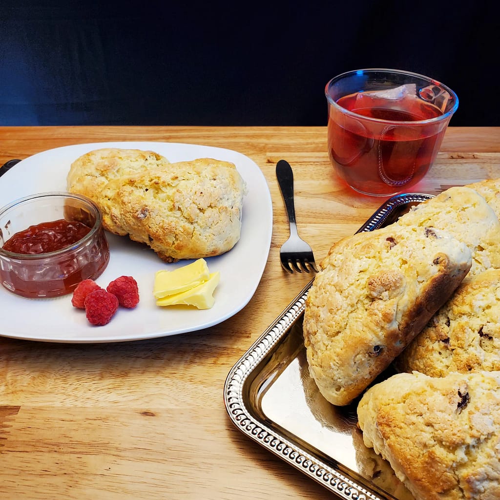 This image depicts cranberry scones in a pile on a silver plater and some serves on a white plate a lot with the raspberry jam, butter and raspberries.