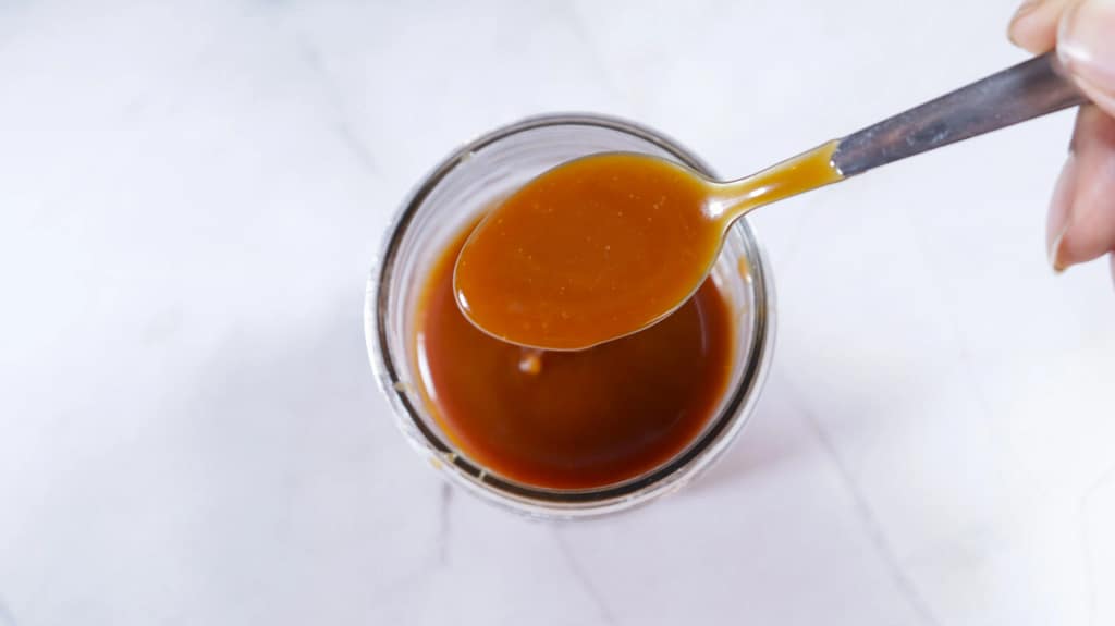 This image shows a glass mason jar filled with caramel sauce with a spoon with some of the caramel sauce.