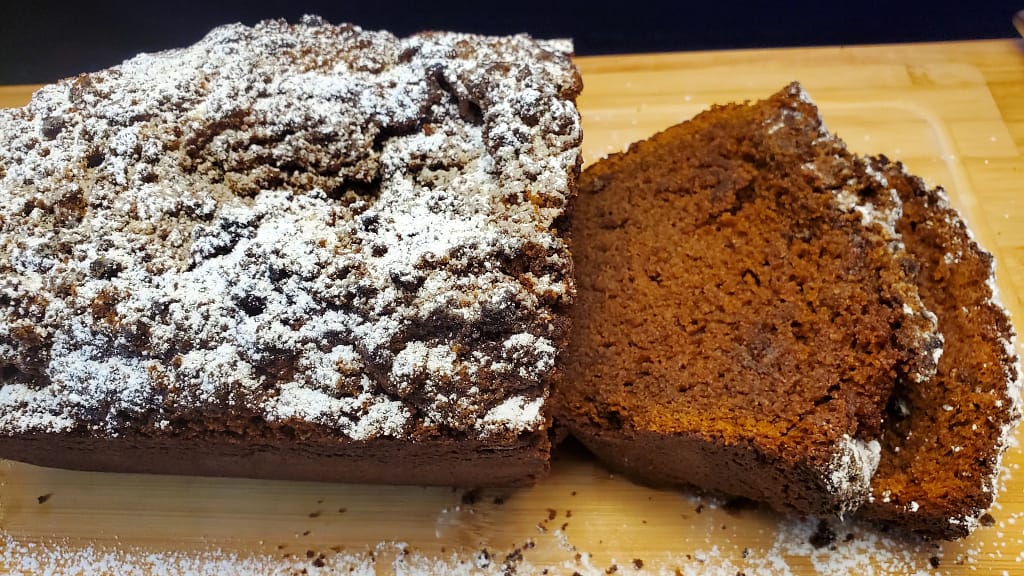 This image depicts the moist chai pumpkin bread as described by this blog post. It is on a wooden cutting board, dusted with powdered sugar and has been sliced twice.