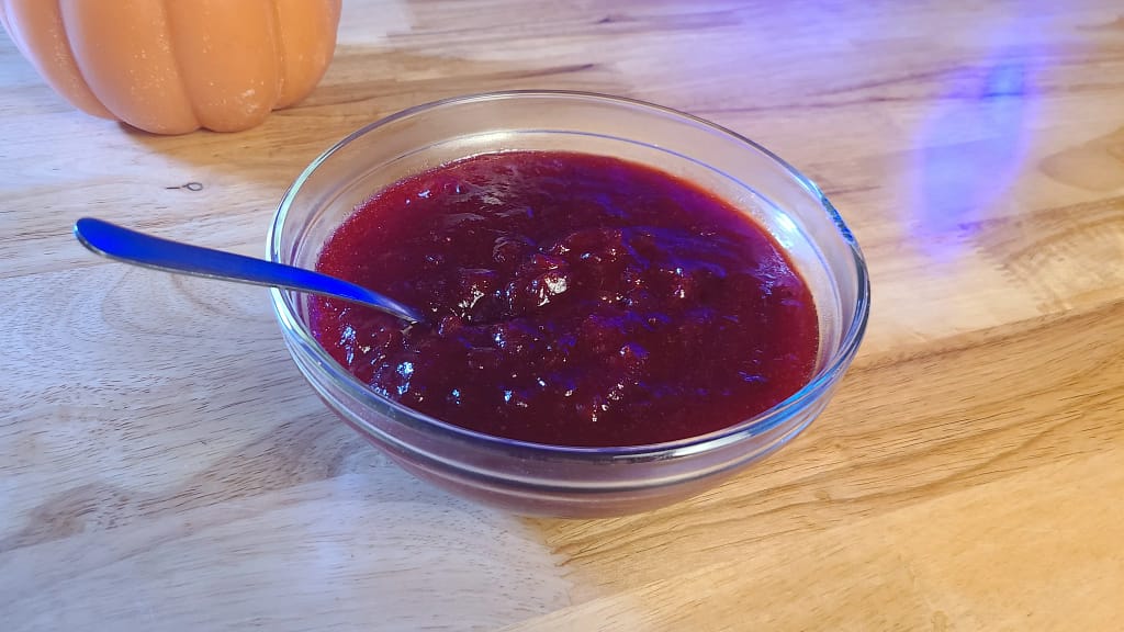 This image depicts the results of following the cranberry sauce recipe on lesliecooks.com. Cranberry sauce is in a clear bowl on a wooden table. A wax pumpkin is the background.