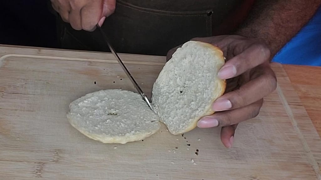This picture depicts a everything bagel being sliced in half on a cutting board.