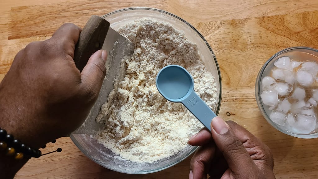 This image depicts a bowl of pie crust ingredients in which iced water is being pour in gradually.