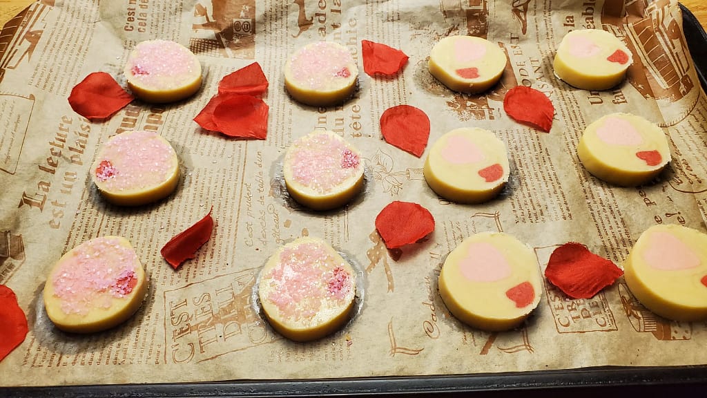 This image depicts unbaked cookies feature in this recipe blog. They are round cookies with heart shaped designs. They are feature on brown parchment paper with artificial rose petals.