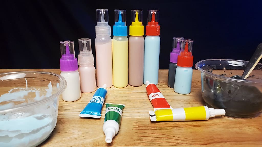 This image shows bottles of royal icing colored different colors. From left to right, white, pink, yellow, purple, blue, black, and blue. On the table there is blue, green, red, and yellow gel food coloings.