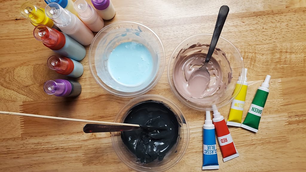 This image shows royal icing colored black, blue, and purple. There are different colored royal icing in bottles. There are also tubes of blue, red, yellow, and green gel food coloring. They are all on a brown wooden table.