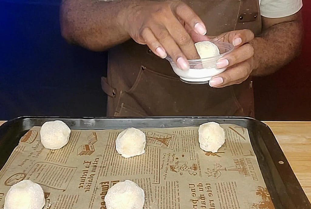 This image depicts Jumbo sugar cookie dough being rolled in white granulated sugar. Dough balls are placed on cookie sheet lined with decorative parchment paper.