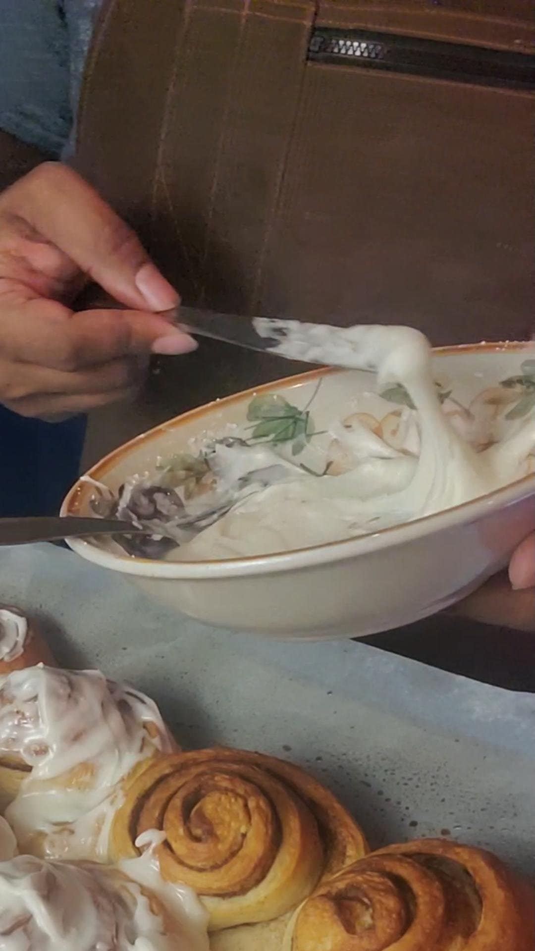 This image depicts a bowl of cinnamon roll icing being spread on soft yeast dough cinnamon rolls as described by the recipe found on lesliecooks.com.