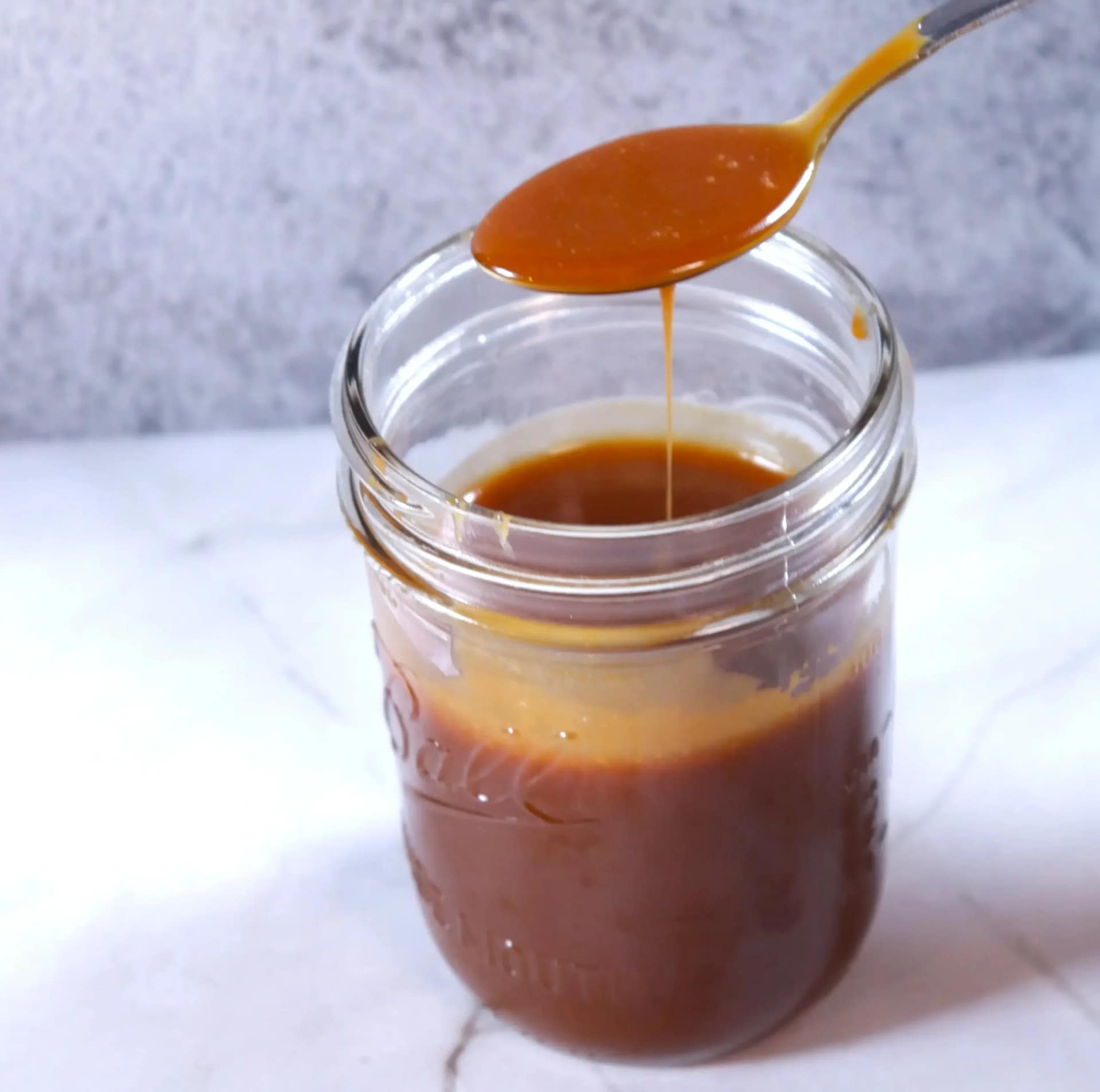 This image shows a glass mason jar filled with caramel sauce with a spoon with some of the caramel sauce.