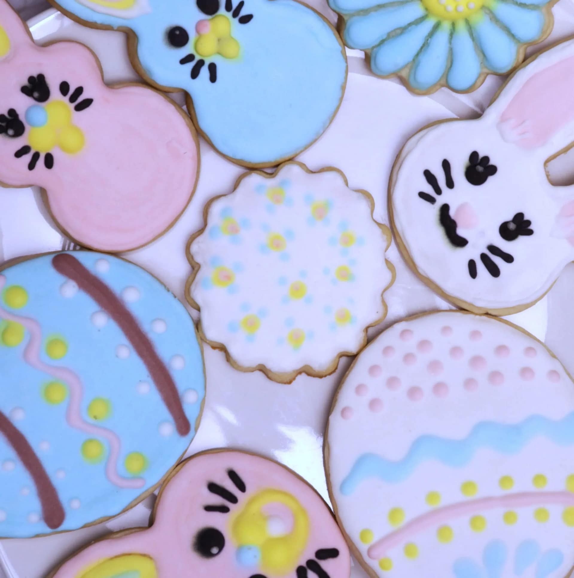 This image show painted sugar cookies on a white plate. There a decorated bunny shapes and egg shapes. They are pink, blue, and white with yellow accents.