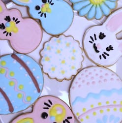 This image show painted sugar cookies on a white plate. There a decorated bunny shapes and egg shapes. They are pink, blue, and white with yellow accents.