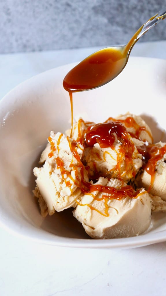 This image shows some caramel sauce being spooned over some black walnut ice cream in a white bowl.