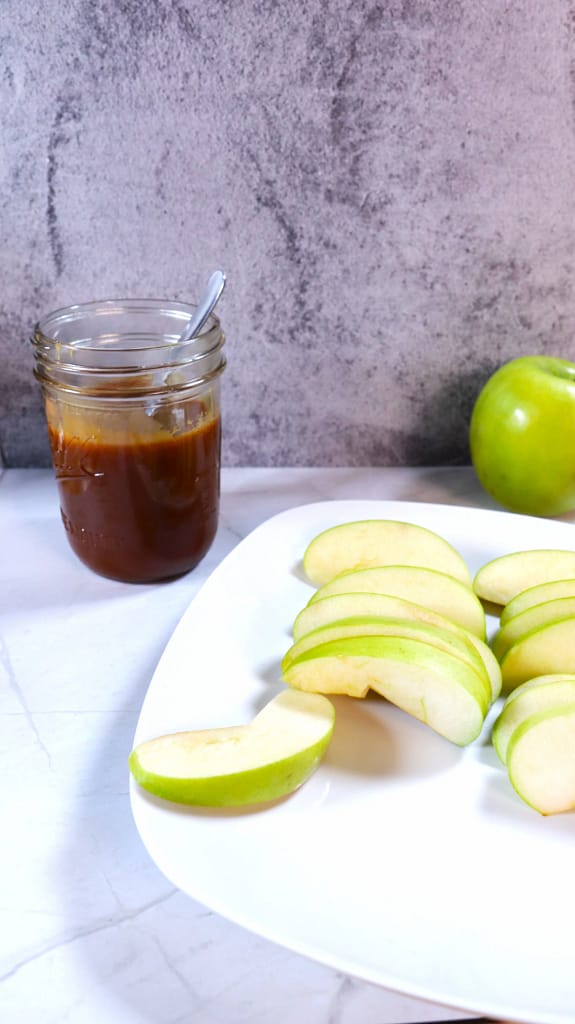 This image shows caramel sauce in a mason jar. A plate of sliced green apples is on the right along with a whole green apple.