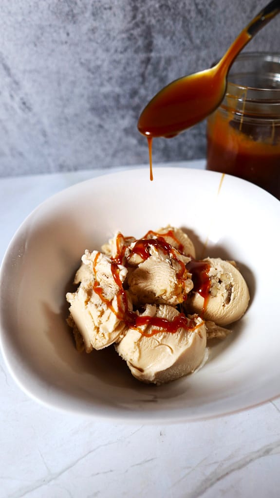 This image shows some caramel sauce being spooned over some black walnut ice cream in a white bowl.