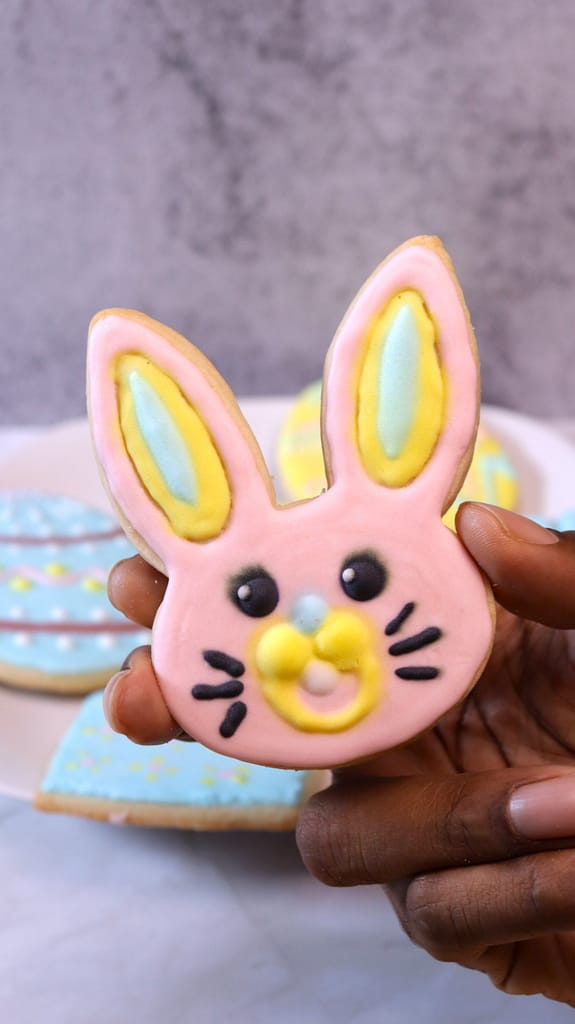 This image shows a pink bunny cookie being held. Blue painted cookies are in the background on a white plate.