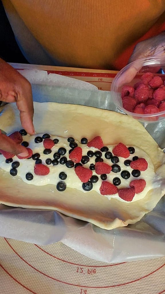 This image depicts blueberries and raspberries being put on top of cream cheese filling. The filling is spread over yeast dough as described by recipe on lesliecooks.com.