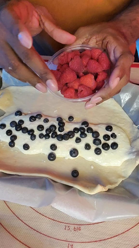 This image depicts blueberries put on top of cream cheese filling. Raspberries are in a bowl. The filling is spread over yeast dough as described by recipe on lesliecooks.com.