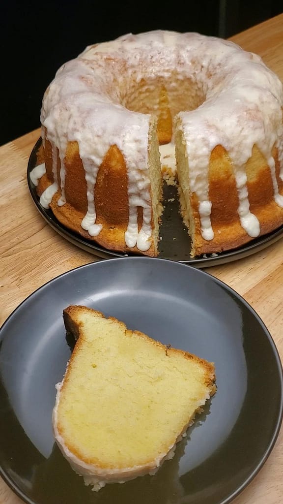 This image depicts 5 flavor pound cake as described by this blog post being sliced. The cake is a nice pale yellow color, The cake is covered with a white lemon glaze. A slice of cake is placed on a black plate.