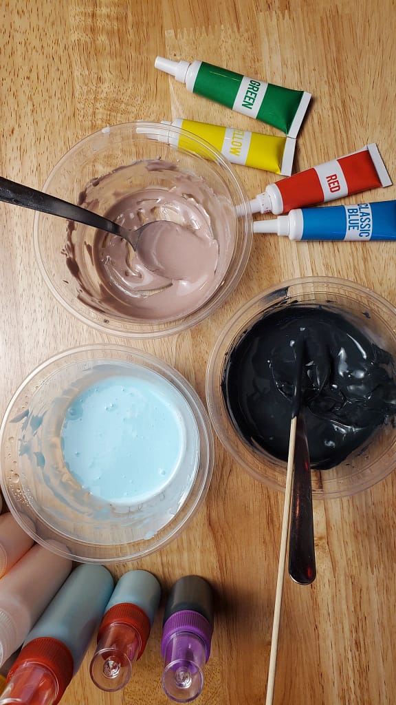 This image shows bowls of royal icing being mixed. There is black, blue, and purple. There are tube of gel food coloring on the brown wooden table. There are also bottles of different colored royal icing.