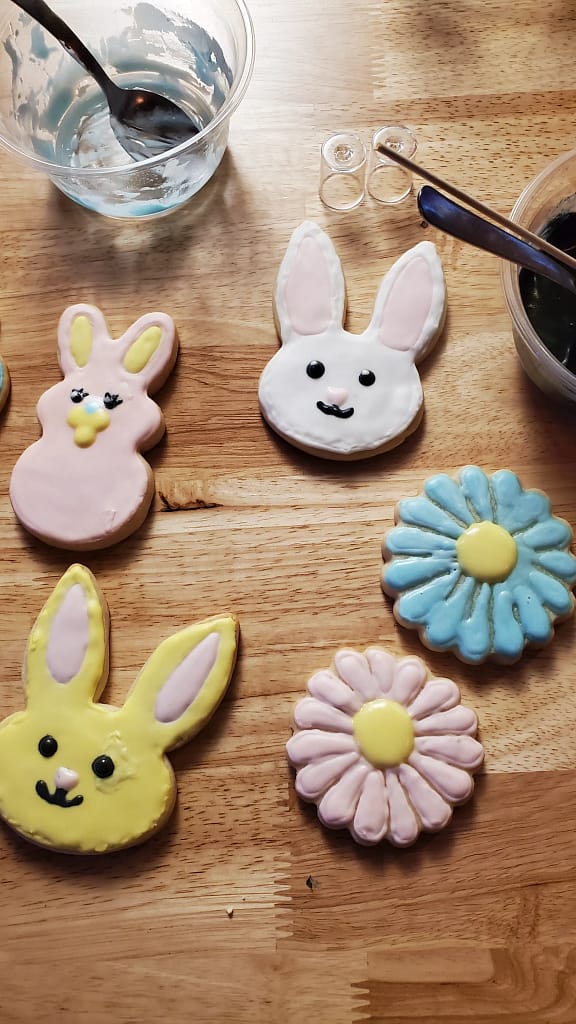 This image show some roughly painted sugar cookies. There are pink and blue flower cookies. There is a pink, yellow, and white bunny cookie.