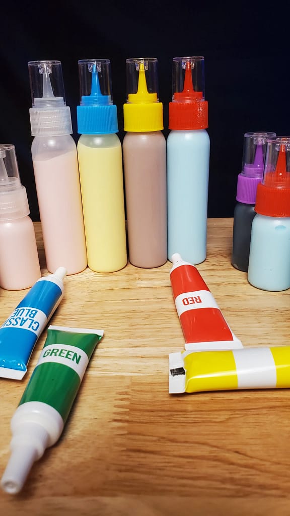 This image shows piping bottles filled with royal icing colored pink, yellow, purple, and blue. There are gel food coloring laying on the table.