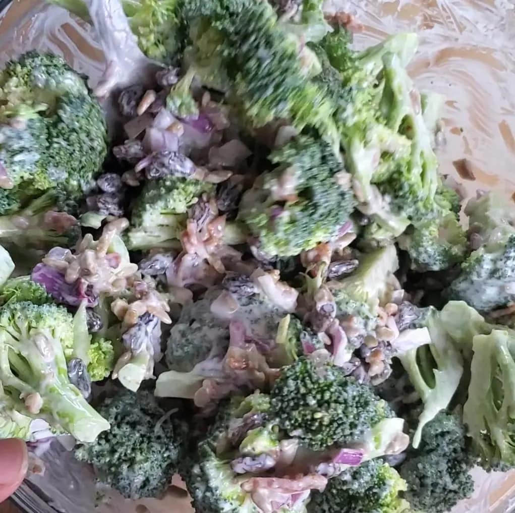 This image depicts easy broccoli salad mixed together in a clear glass bowl. You can see the green broccoli, purple raisins, and bacon all coated in a creamy dressing.
