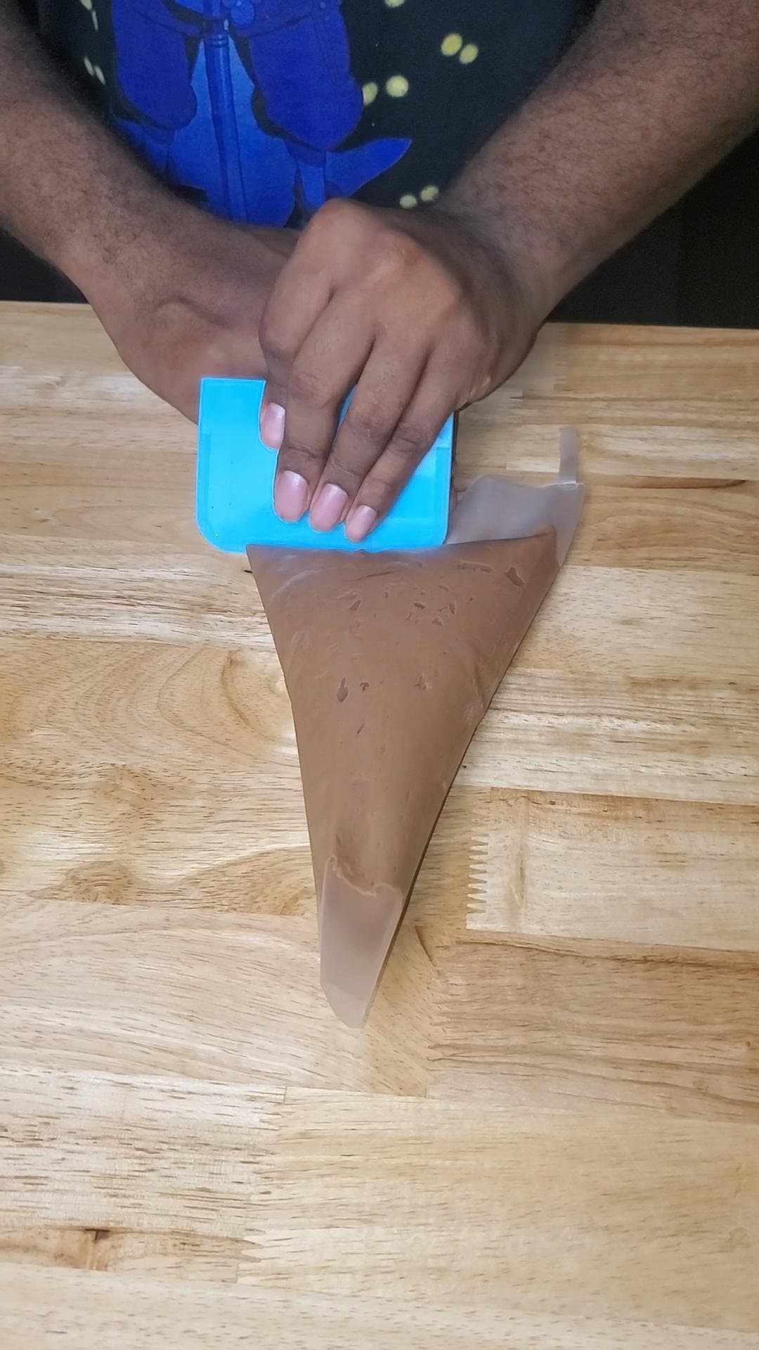 This image show chocolate cream cheese frosting being pushed down into a piping bag using a blue bench scraper.