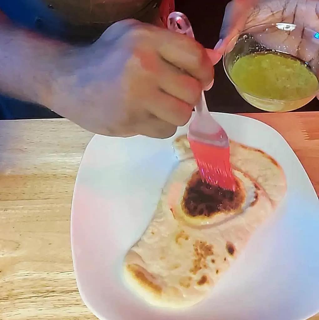 This image depicts a cooked piece of naan being spread with garlic butter using a red silicone brush.