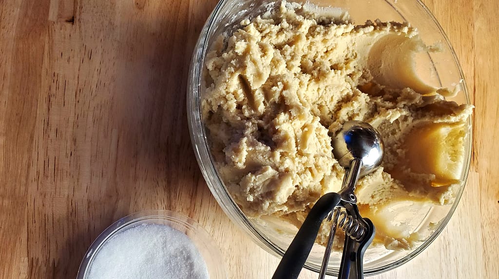 This image depicts a large glass bowl of Jumbo Sugar Cookie dough. There is a small bowl of white sugar to lower left of the cookie dough.