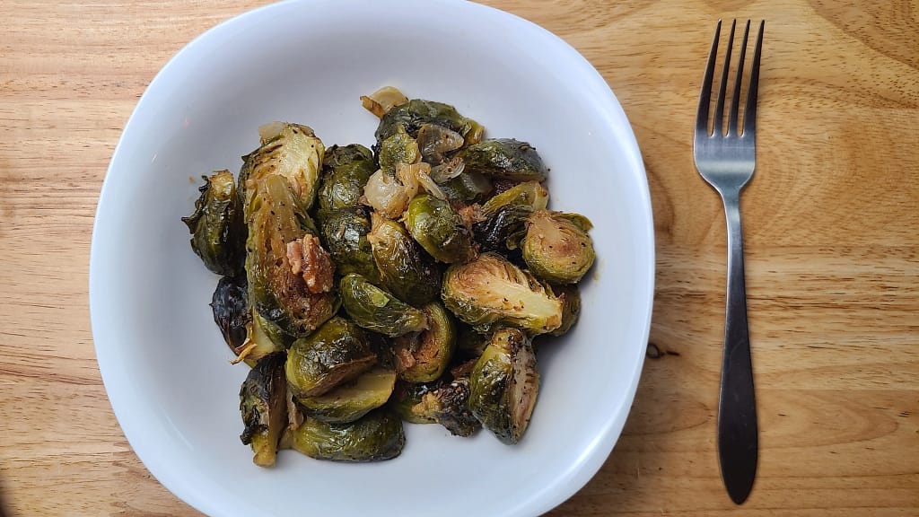 This image depicts the roasted brussels sprouts in a white bowl and a fork to the right of the bowl. They are on a wooden table.