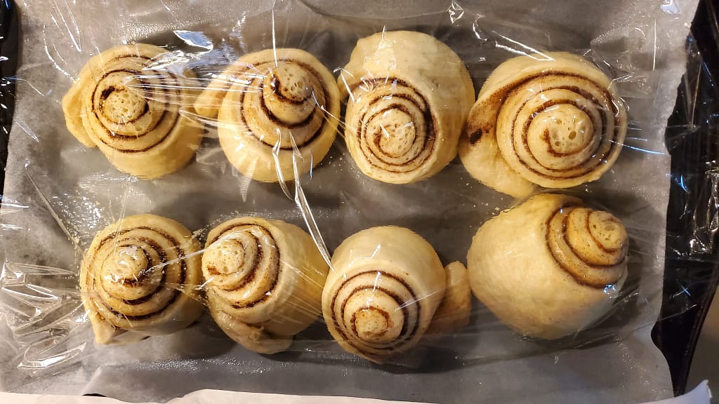 This image depicts Always soft cinnamons shaped and going through second proofing. They are on a cookie sheet, lined with parchment paper, and covered with greased plastic wrap.