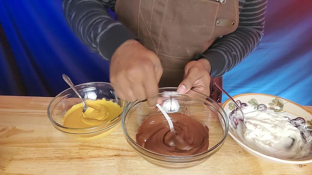 This image depicts Leslie stirring 3 bowls of chocolate. They are butterscotch, semi-sweet chocolate, and white chocolate starting from left to right.